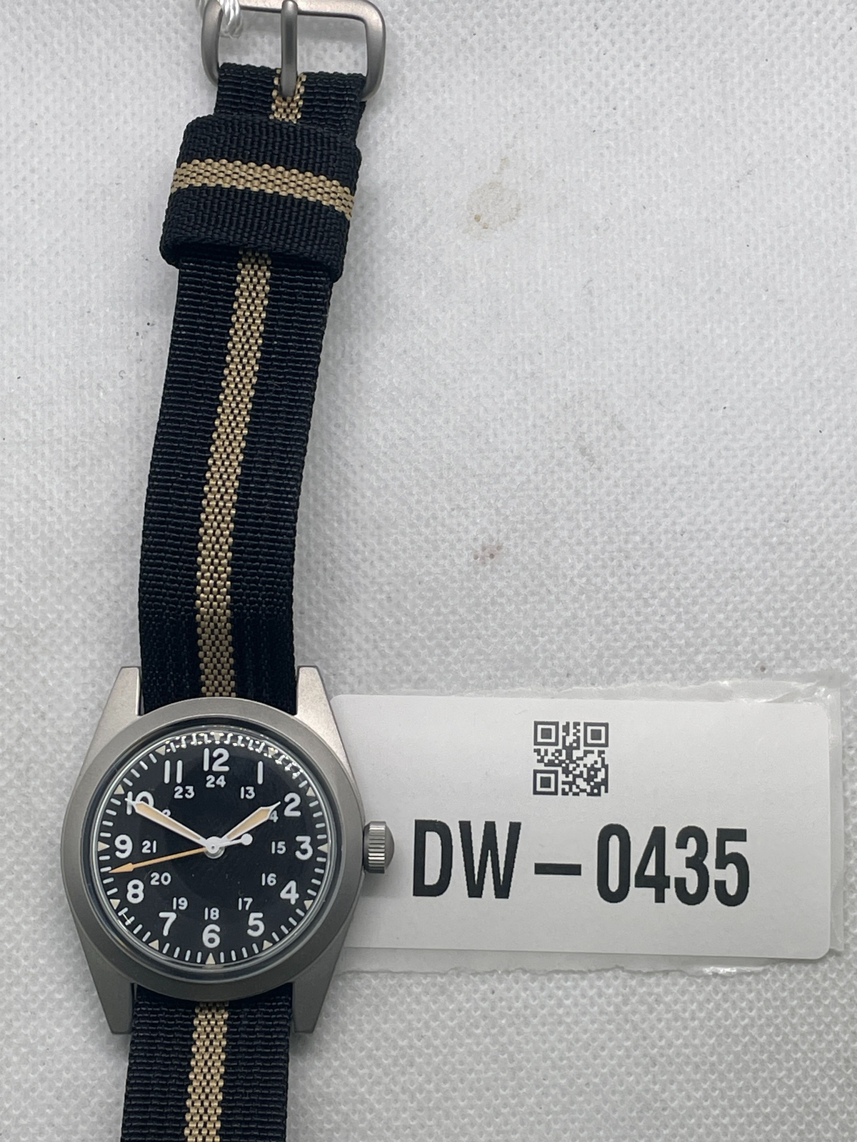 Pre-owned Rdunae field watch with a black 12- and 24-hour dial and black fabric strap with a tan stripe, beside tag DW-0435.